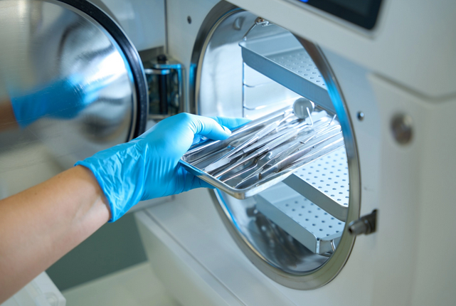 Hand mit blauer Schutzhandschuhen legt ein Metalltablett mit medizinischen Instrumenten in einen Autoklaven zur Dampfsterilisation – Beispiel für hygienische Aufbereitung bei 121